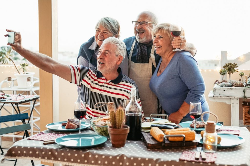 Happy seniors friends taking selfie with mobile smartphone camera at dinner on terrace - Retired people having fun eating and drinking red wine on patio while using new trends technology phone apps I StrongLink Technology