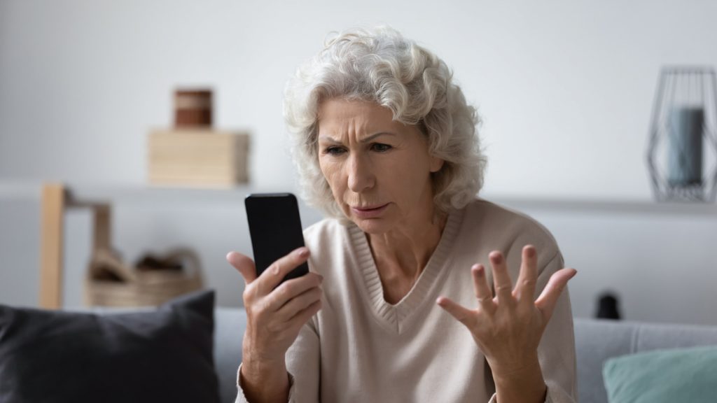 Angry mature woman sit on couch at home feel frustrated by slow internet connection on cellphone, mad elderly female confused stressed by smartphone operation problems, virus or spam messages